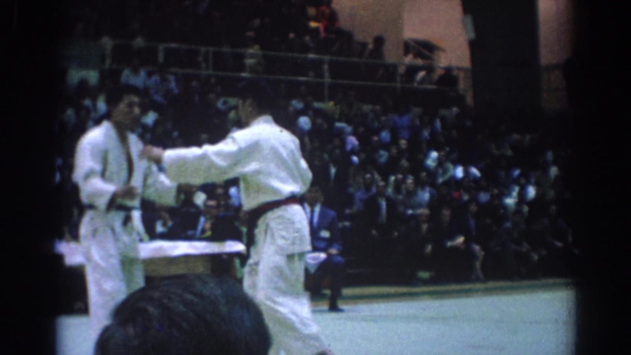 Two men in white gi face each other on a gym floor before crowded bleachers.