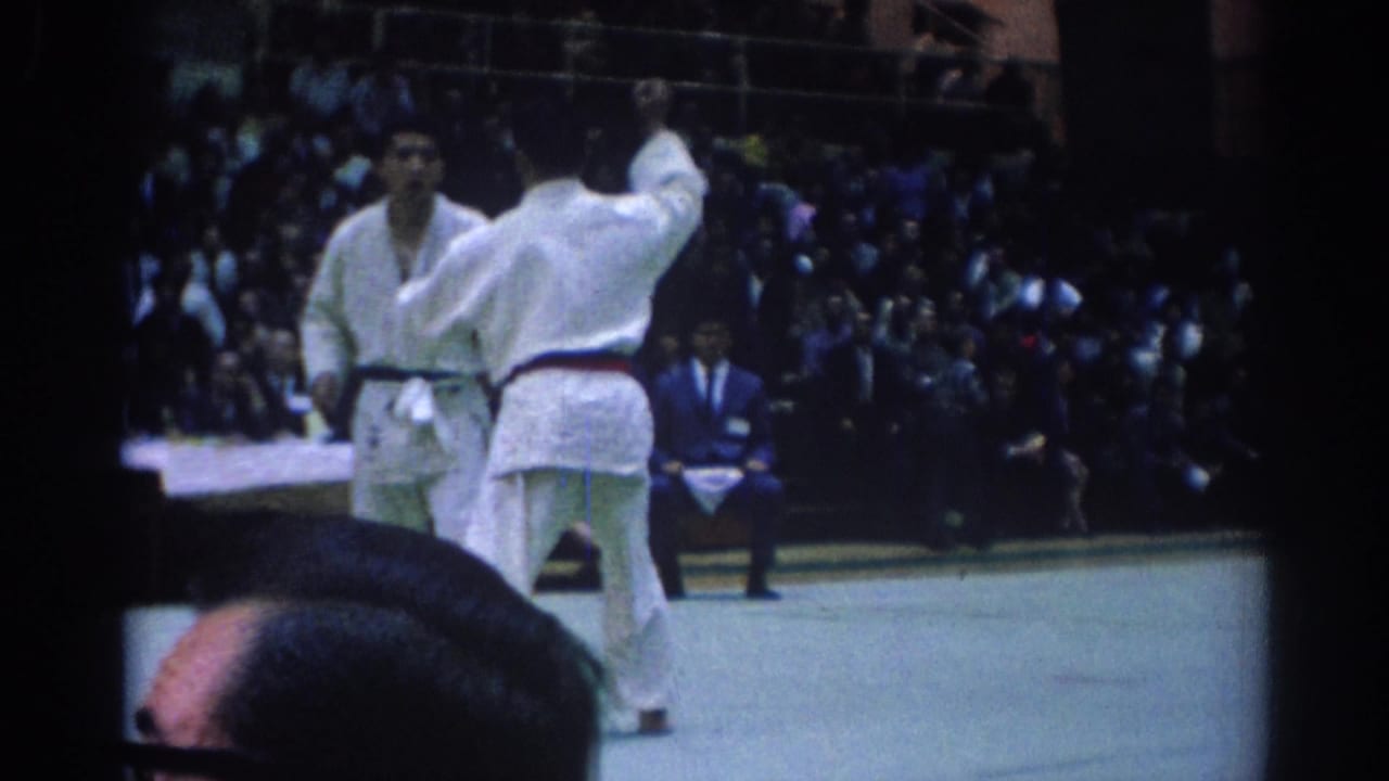 A martial arts competitor raises an arm while facing another black-belt competitor in the 1967 Los Angeles gymnasium footage.