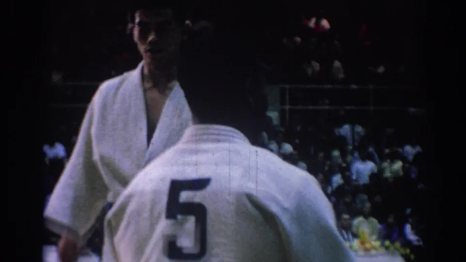 A numbered competitor in a white gi stands foreground while another martial artist faces him in the crowded gymnasium.