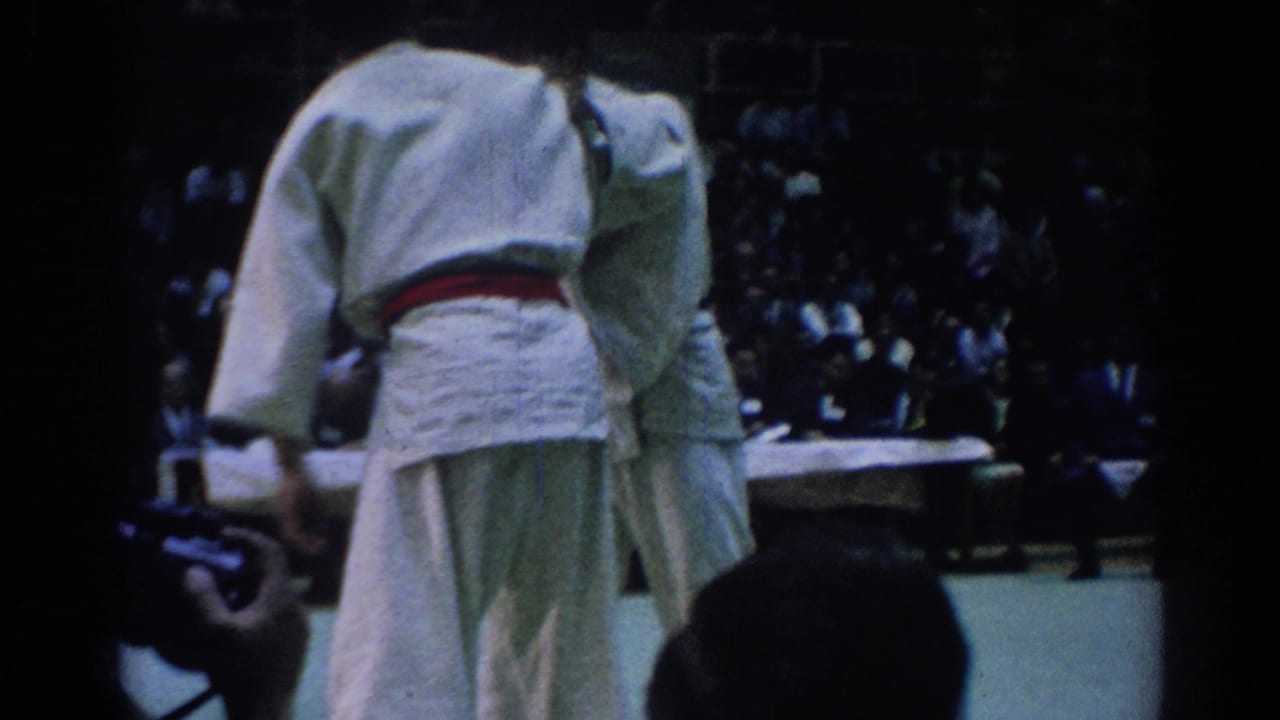 Judo athlete in a white gi with a red belt stands near the match mat and crowd.