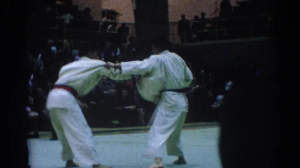 Two judo competitors in white gi and colored belts lean into a grip exchange on the 1967 Los Angeles arena mat.