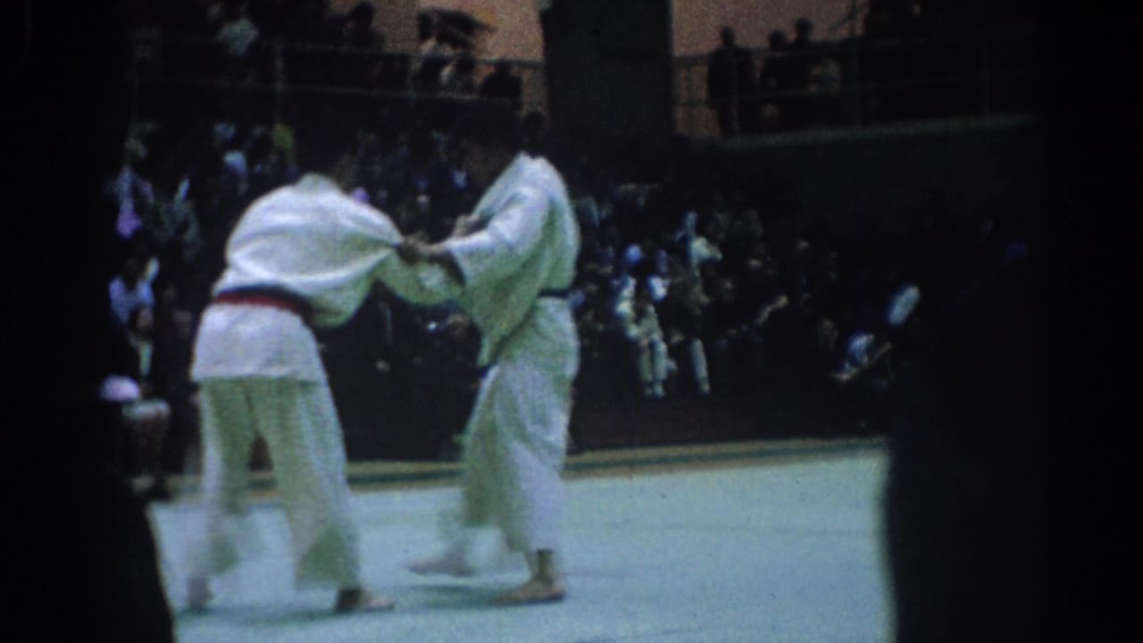 Two judo athletes in white gi grapple on a mat before dark spectator seating.