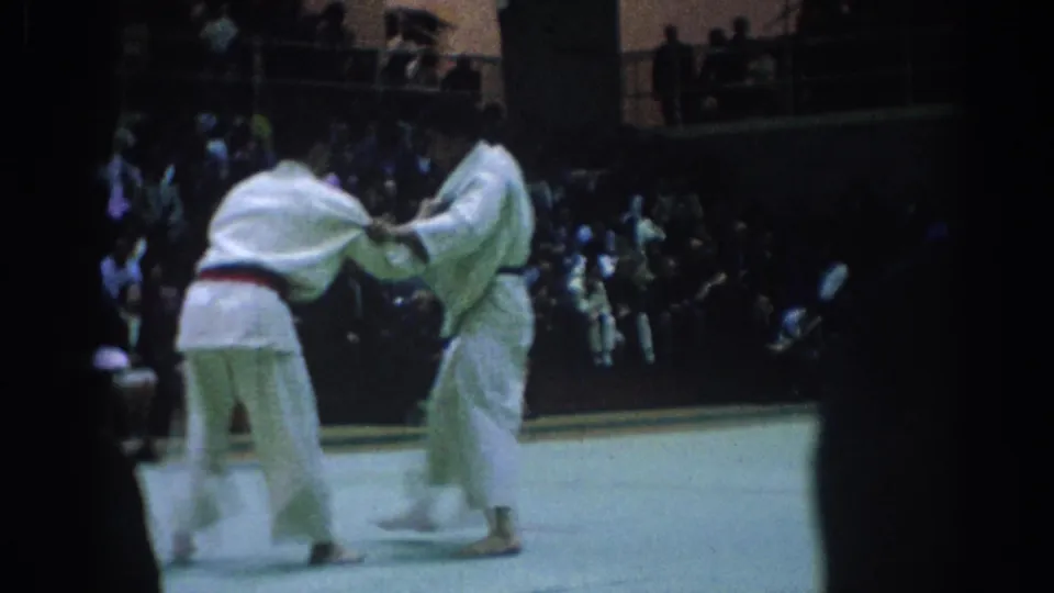 1967 Los Angeles 8mm frame showing two judo athletes in white gi gripping each other during an indoor match before seated spectators.