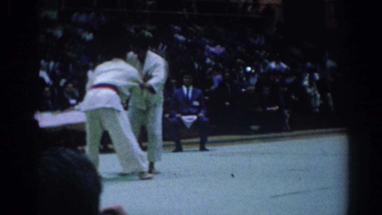 Judo competitors lean into a grip exchange on the mat with an official nearby.