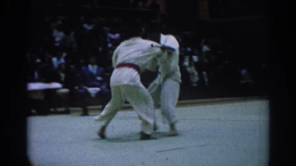 1967 Los Angeles 8mm frame showing a judo athlete in a red belt bending into a throw attempt against an opponent in white gi.