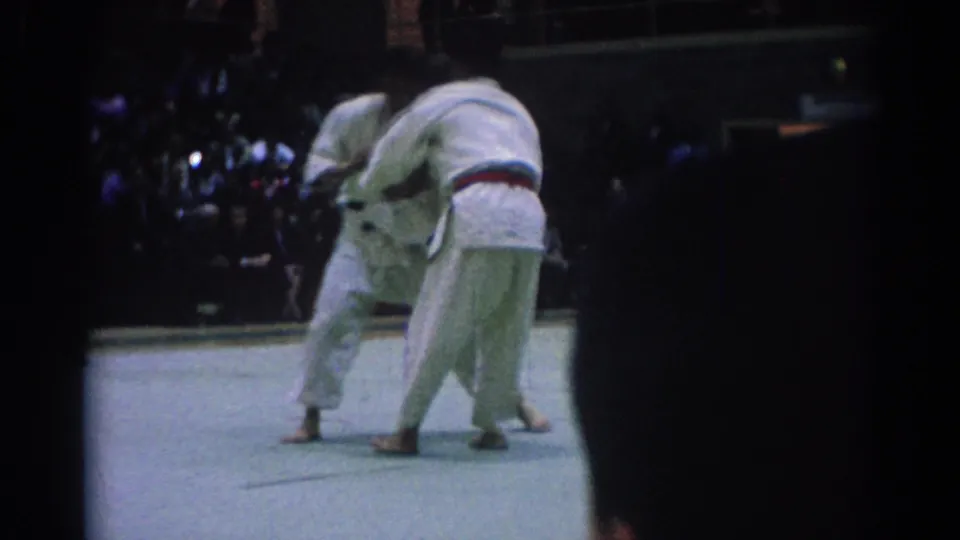 1967 Los Angeles judo competition frame with two athletes in white gi locked in close contact before a seated audience.