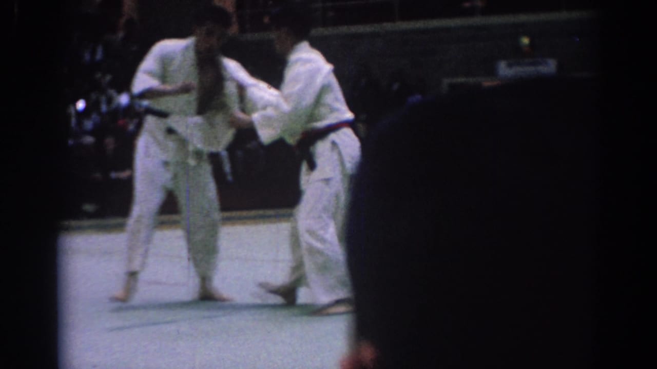 Two men in white gi spar on a gym floor while spectators sit in the background.