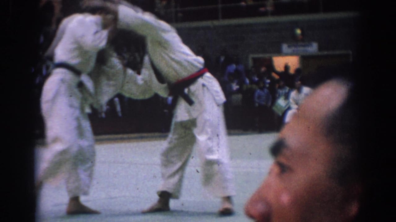 1967 Los Angeles home-movie frame of two gi-clad men in close sparring contact with a foreground spectator visible.