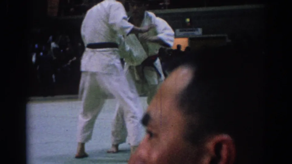 1967 Los Angeles 8mm frame with one martial artist upright and another bent low, seen from the audience area.