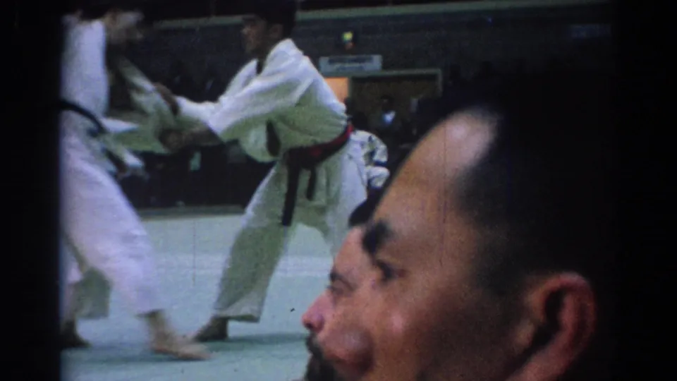 1967 Los Angeles home-movie frame of a red-belt martial artist crouching during sparring, with a spectator face in front.