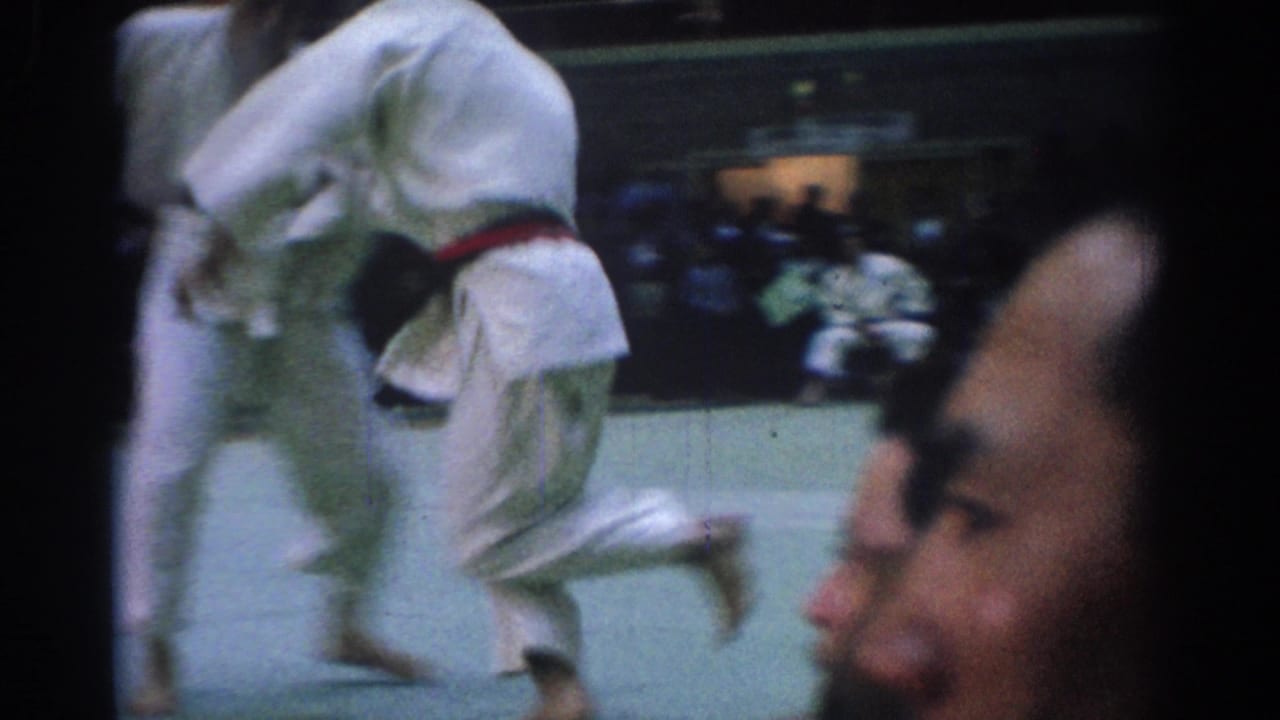 Two judo athletes in white gi move across a mat as spectators sit behind them.