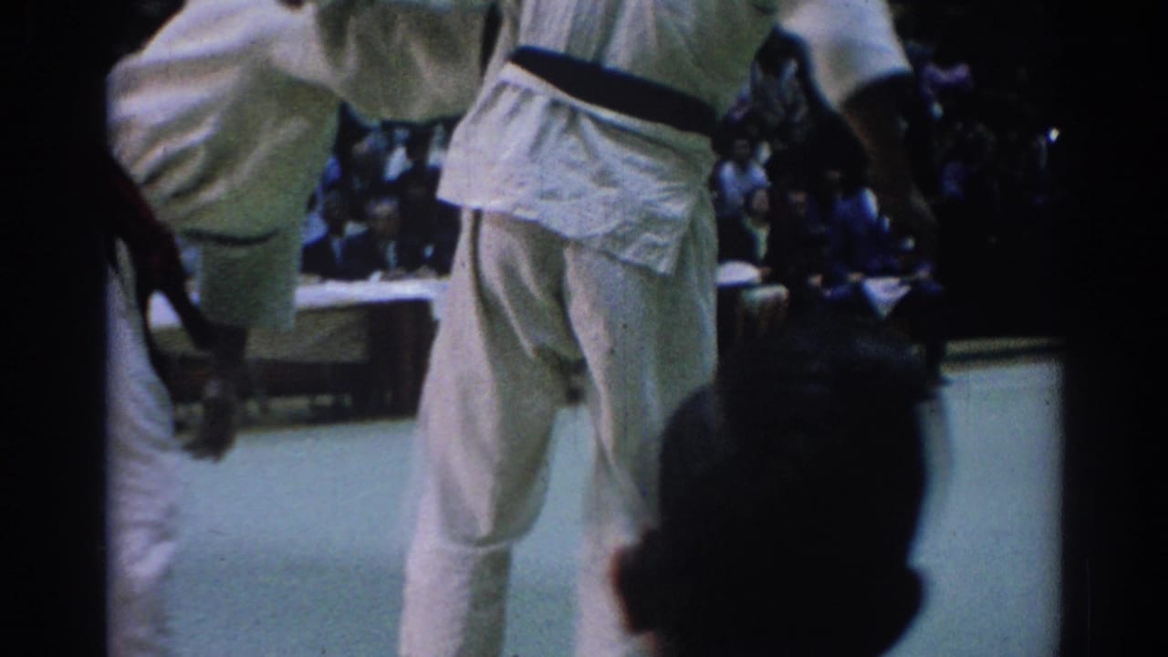 A 1967 Los Angeles judo match continues on a pale indoor mat, with competitors in white gi and a seated audience behind them.