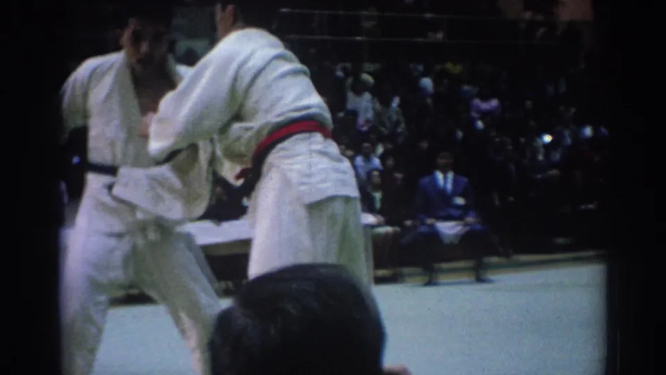 Two judo competitors in white gi grapple during a 1967 Los Angeles indoor competition, with spectators and officials visible beyond the mat.