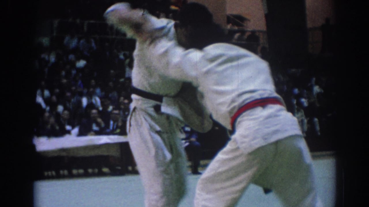 Two judo competitors in white gi grapple near the edge of an indoor mat.