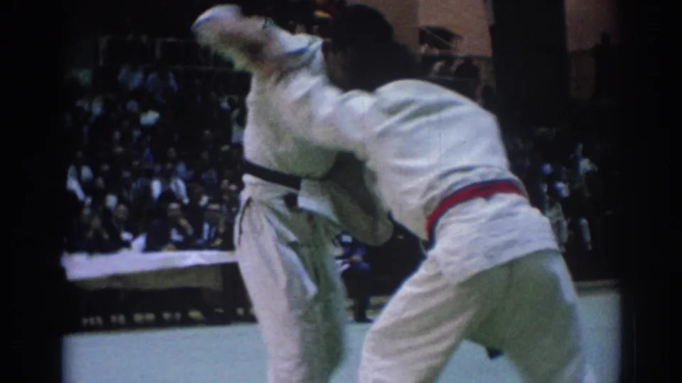 1967 Los Angeles 8mm frame showing two judo competitors in white gi locked in close contact before a seated crowd.