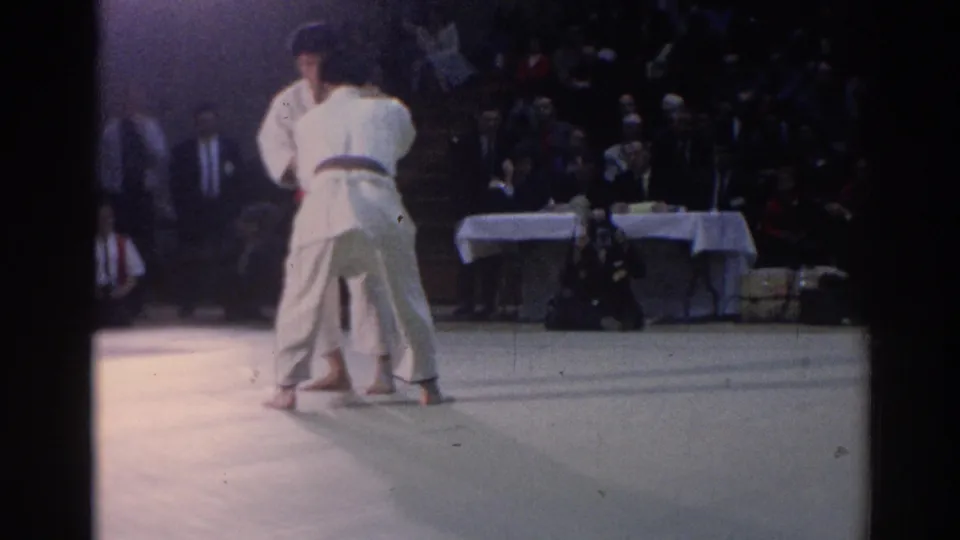1967 Los Angeles frame from behind the competitors, showing a close judo clinch with judges' tables and spectators beyond the mat.