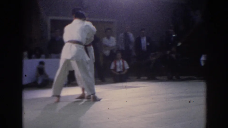 1967 Los Angeles judo match frame showing the athletes locked together under arena lights, with officials and spectators behind them.