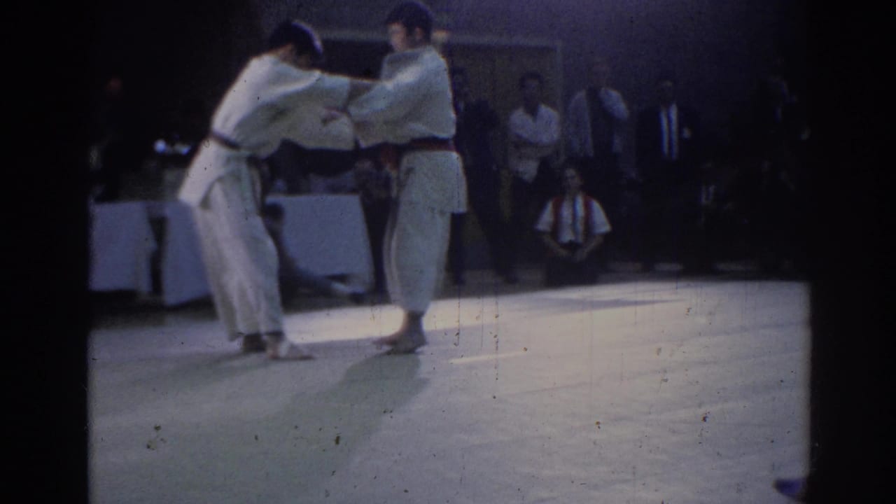 Two judo competitors in white uniforms grapple on a mat before seated spectators.