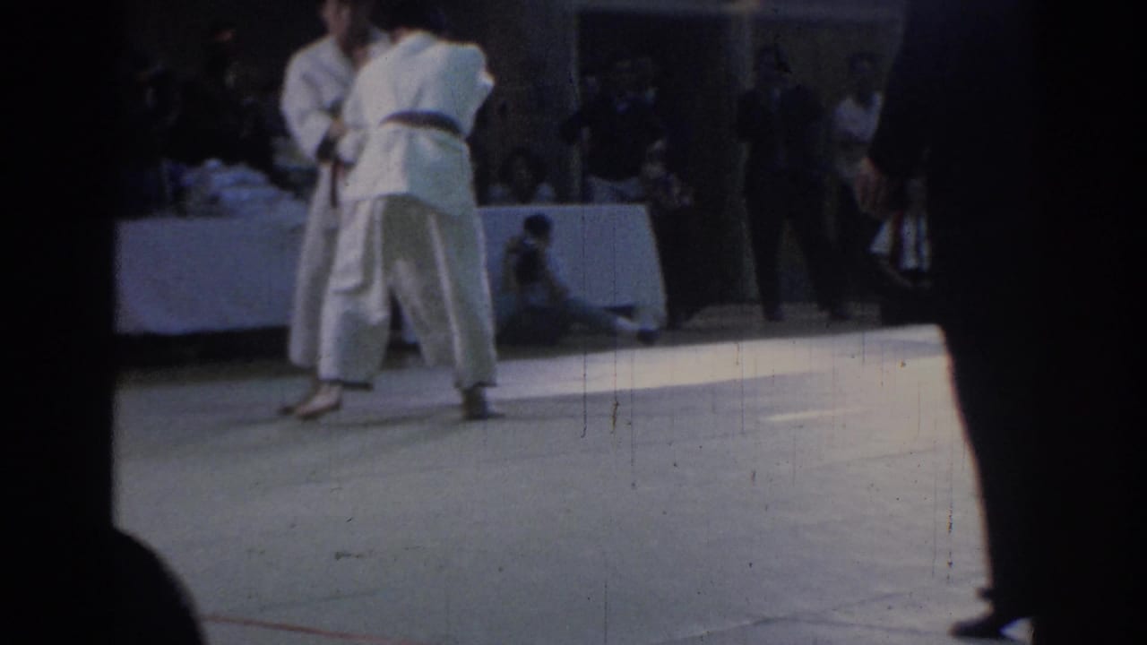 Two competitors in white judogi continue a standing exchange on a light mat in the Los Angeles judo venue.