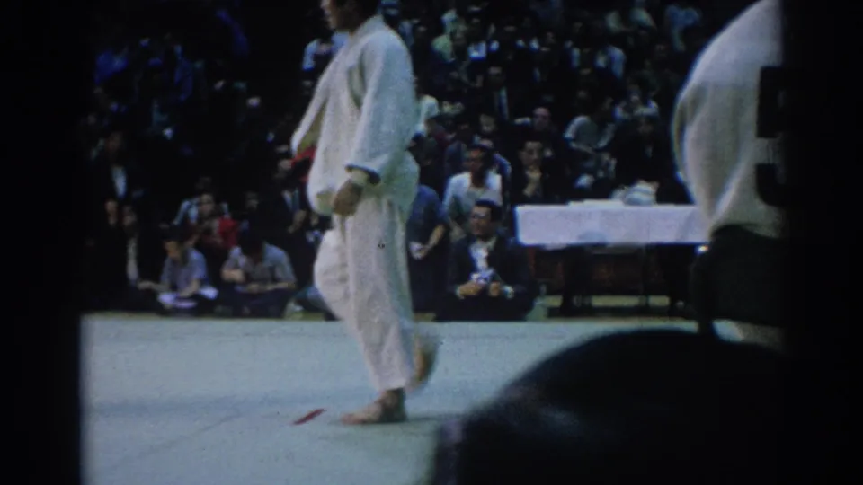 A competitor in a white judogi walks across the mat as the crowd watches from the edge of the 1967 Los Angeles venue.