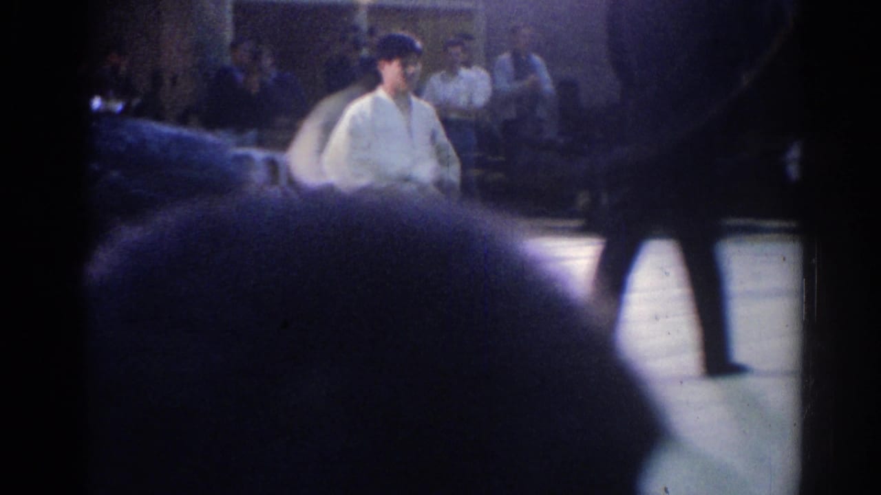 A white-uniformed judo competitor is partly blocked by spectators and a passing figure in the dim 1967 Los Angeles venue.