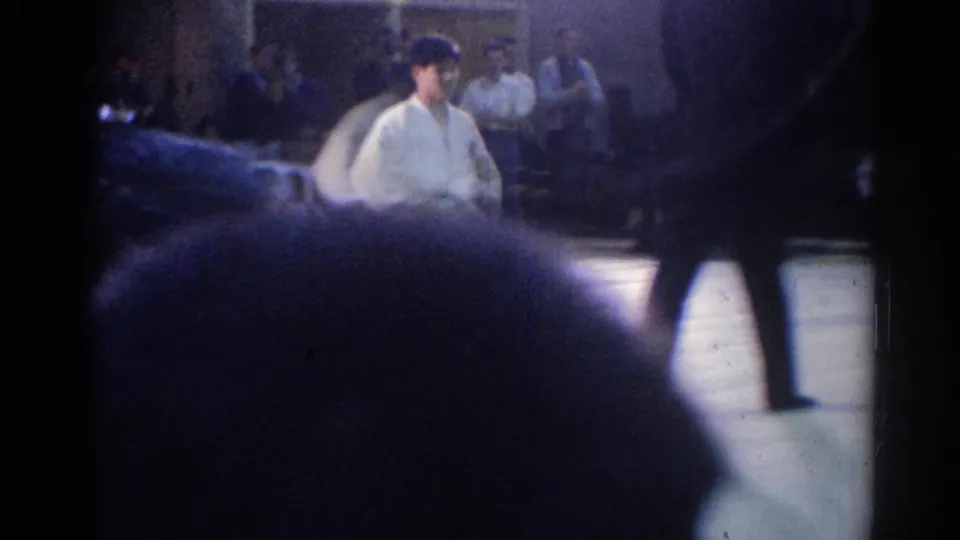 A white-uniformed judo competitor is partly blocked by spectators and a passing figure in the dim 1967 Los Angeles venue.
