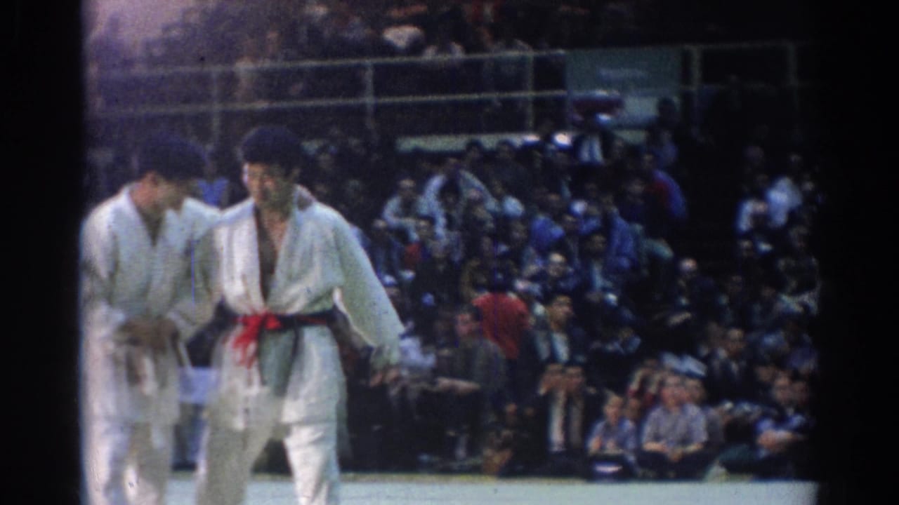 Two men in white gi stand on a mat before a seated audience.
