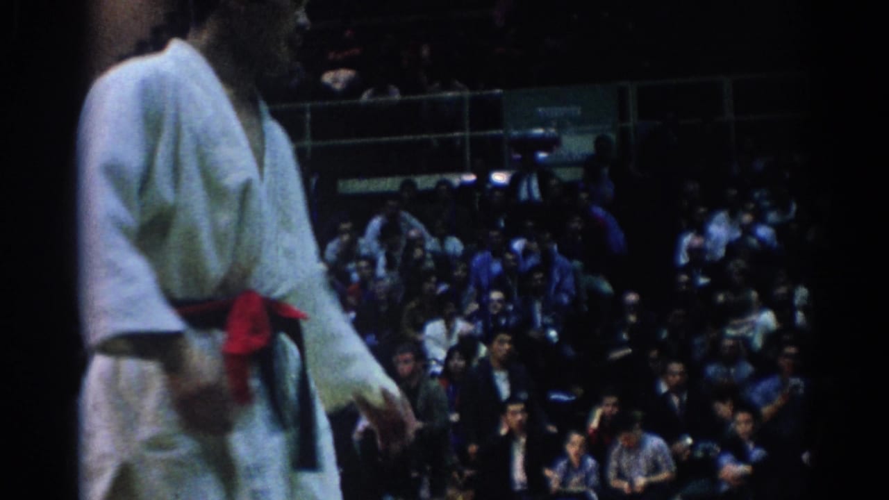 A close, cropped 8mm frame shows a martial arts competitor in a white gi and belt beside the mat with spectators behind him.