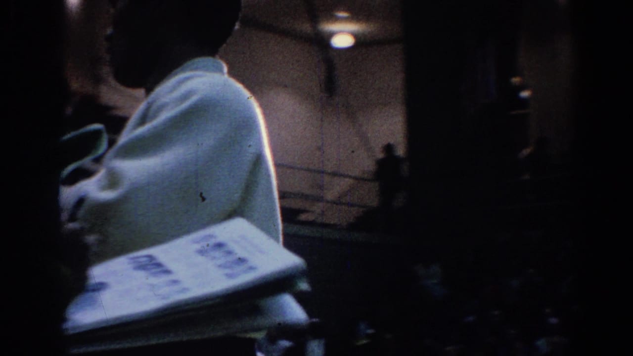 Two white-uniformed martial arts competitors in a dim gym, one partly seen beside a clipboard or papers.