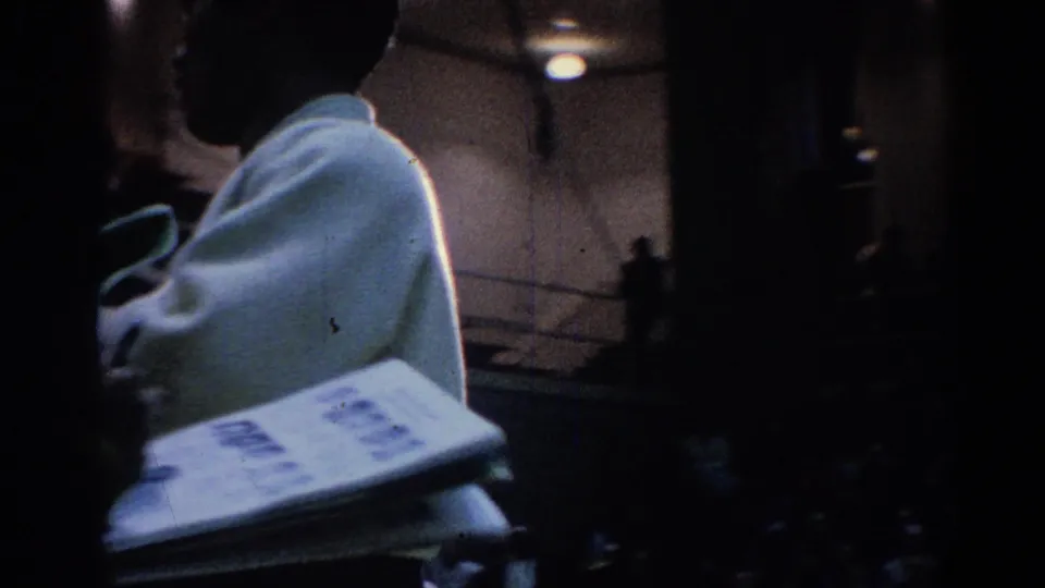 1967 Los Angeles 8mm frame showing martial arts competitors in white gis moving through a dim indoor gym after competition.