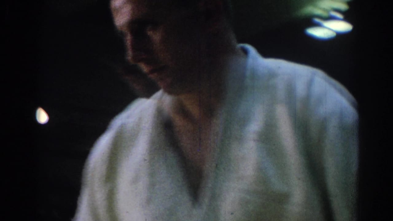 Close 8mm view of an adult martial arts competitor in a white gi walking through a dim Los Angeles gym setting.