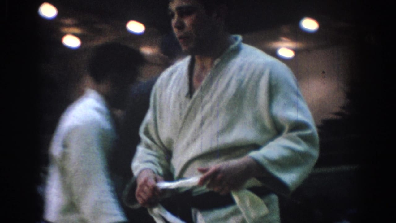 1967 home-movie frame of a white-gi martial arts competitor holding cloth or belt as another competitor walks behind him.