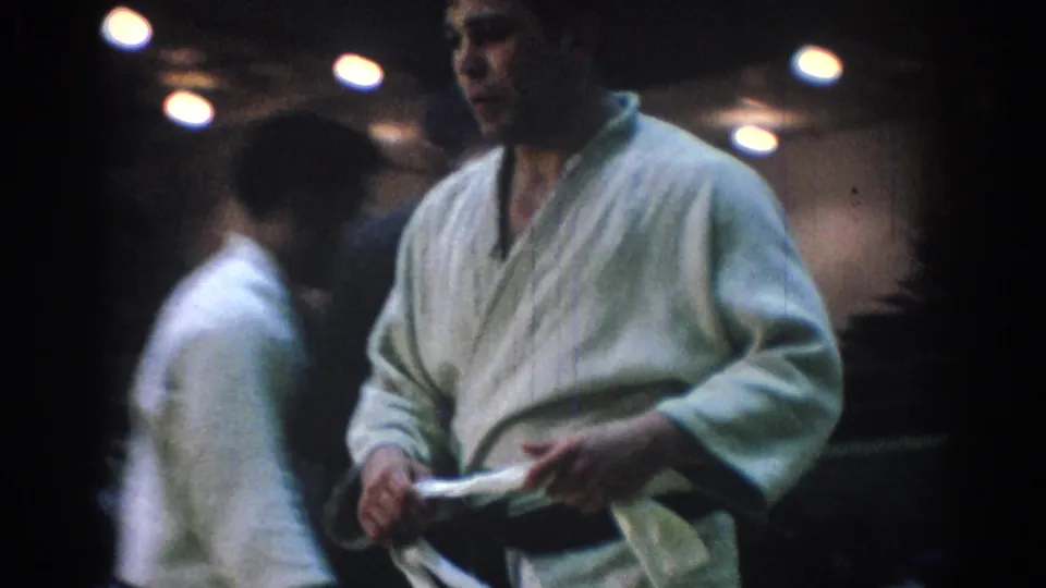 1967 home-movie frame of a white-gi martial arts competitor holding cloth or belt as another competitor walks behind him.