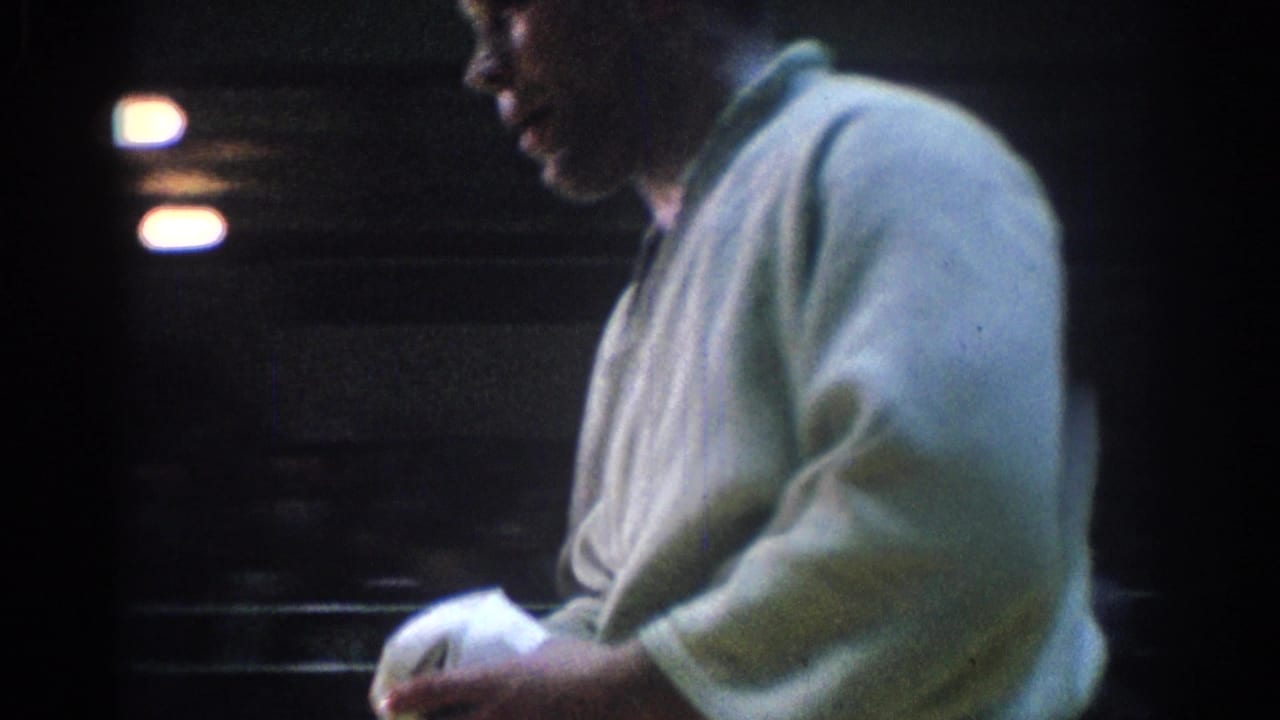 Side-profile 8mm frame of a martial arts competitor in a white gi holding cloth or belt in a dim gym environment.