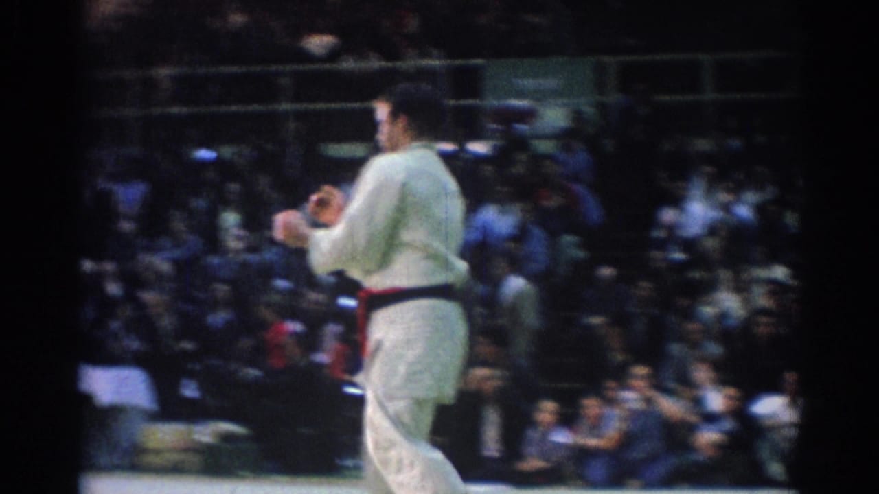 Judo competitor in a white uniform stands on a gym mat with spectators behind him.