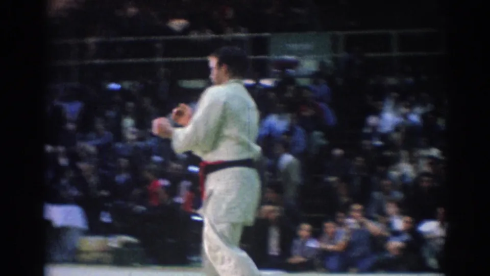 1967 Los Angeles 8mm frame showing a male judo competitor in a white judogi on a mat before seated spectators.