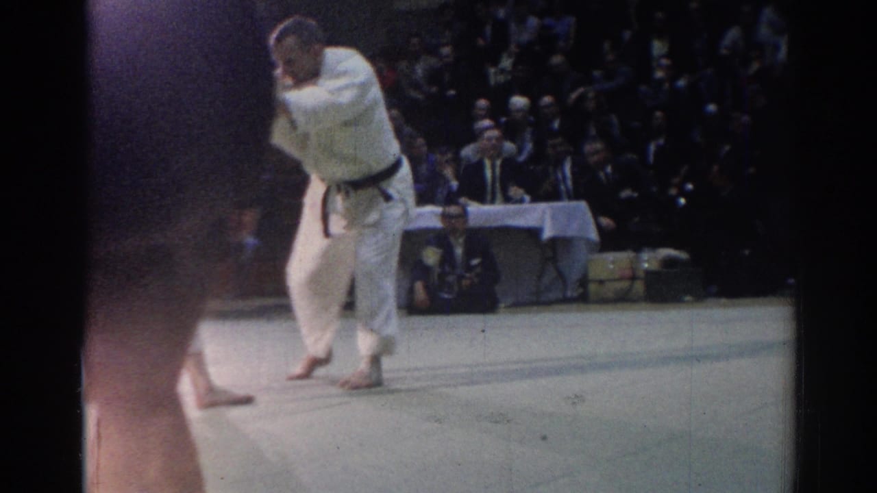 Handheld 8mm view of a judo competitor moving on the mat in a Los Angeles gym, with audience seating behind him.