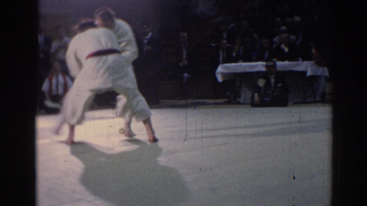 Two judo athletes in white uniforms grapple on a light mat before a dark seated crowd.