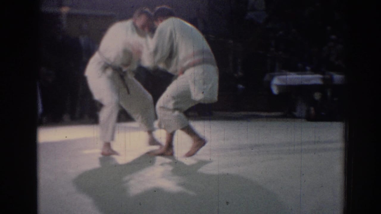Two judo players in white uniforms hold grips and shift their footing on a brightly lit mat in Los Angeles archival footage.