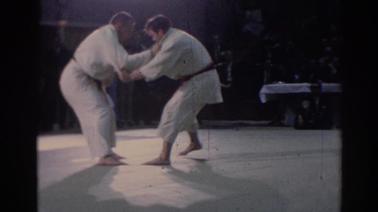 Two judo athletes face off in low stances, with one stepping forward as spectators watch from the dim background.