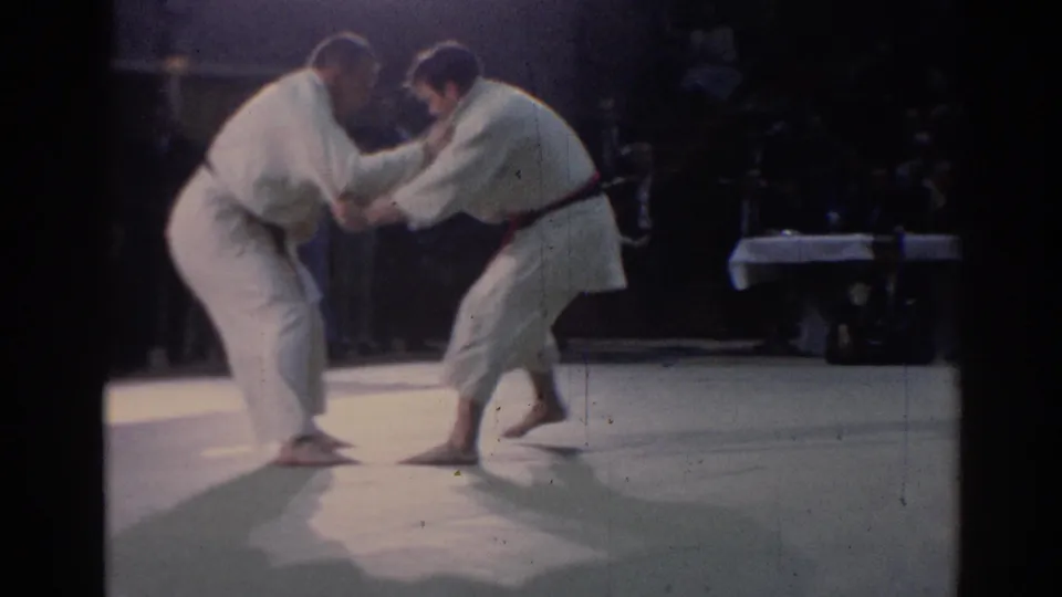 Two judo athletes face off in low stances, with one stepping forward as spectators watch from the dim background.