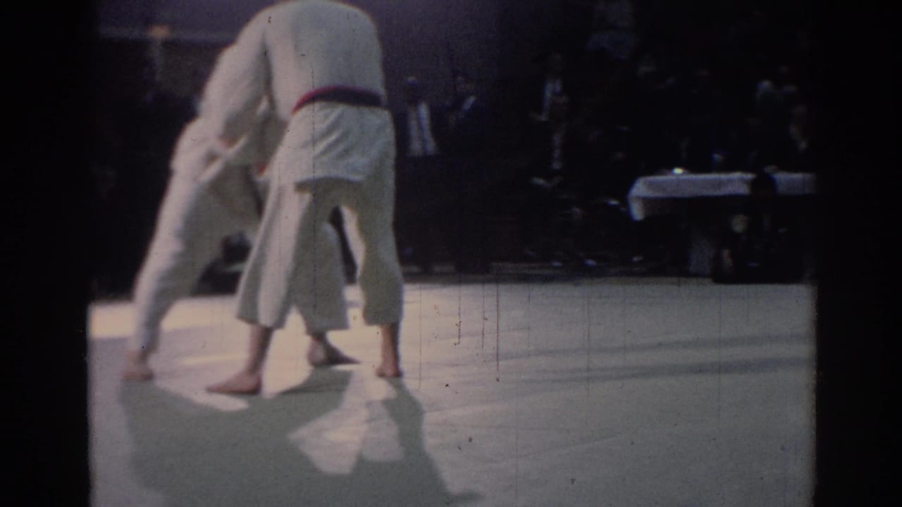 The red-belted judo competitor stands partly back to camera while the other athlete shifts away on the mat.