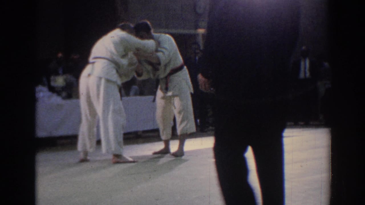 Two judo competitors in white uniforms grapple on a mat near a seated crowd.