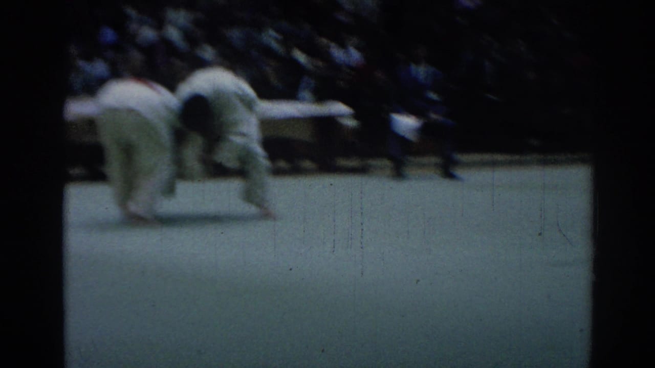 Soft 8mm frame of a judo exchange, with two white-uniformed competitors bent near the mat and spectators in the background.
