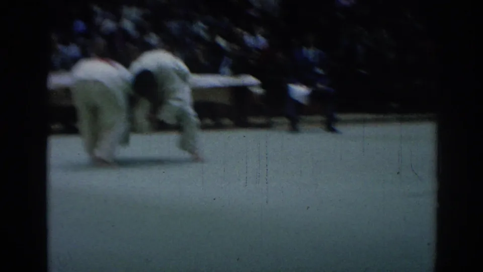 Soft 8mm frame of a judo exchange, with two white-uniformed competitors bent near the mat and spectators in the background.