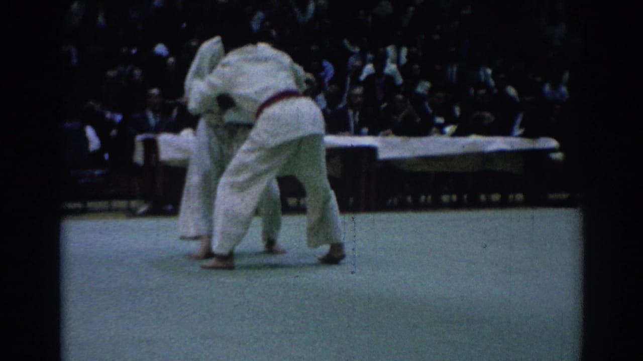 1967 Los Angeles judo competition frame showing white-clad competitors clinched upright, with officials and a crowd behind them.