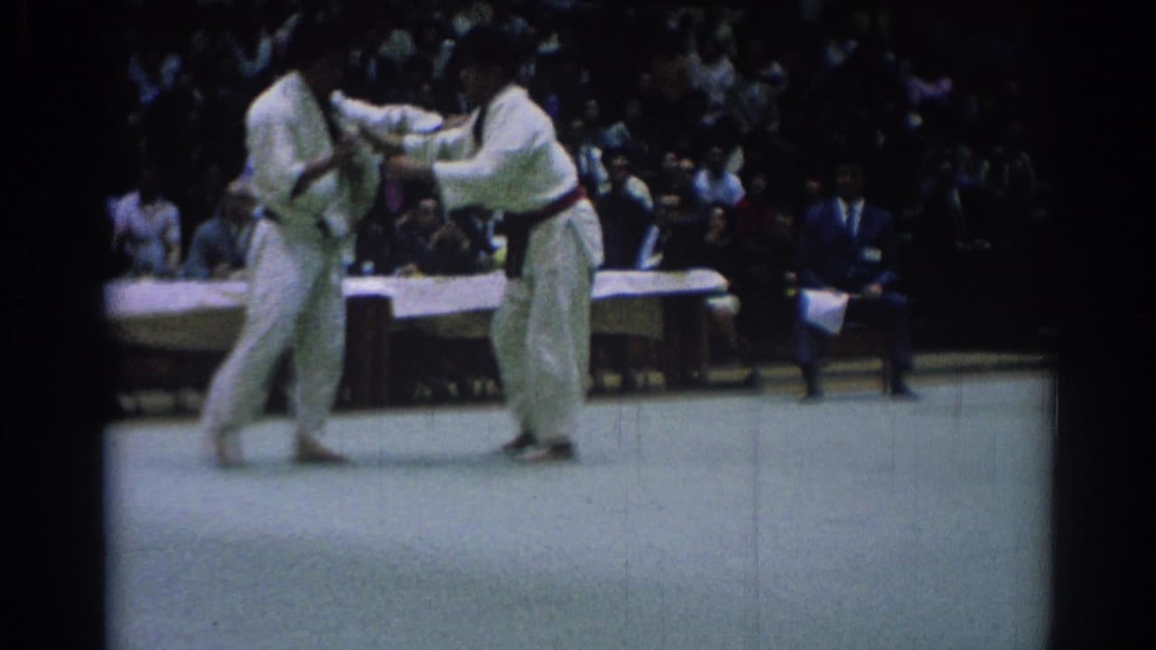 Two karate competitors in white gi grip each other on a mat before seated spectators.