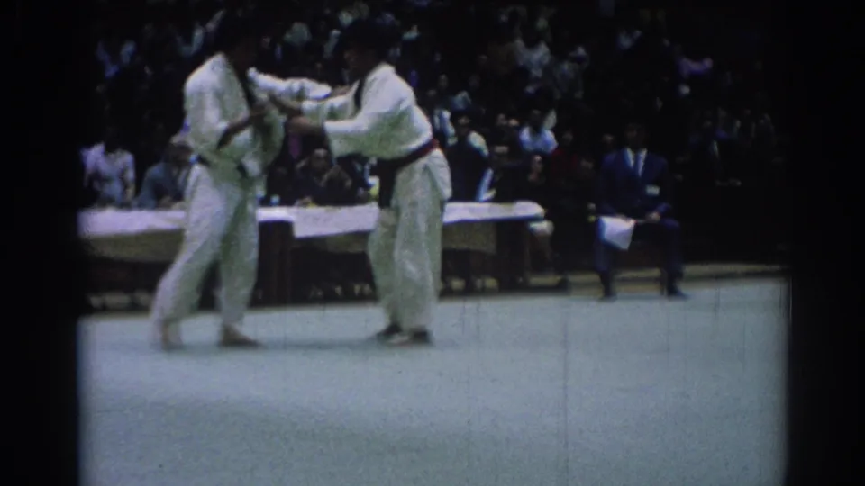 1967 Los Angeles 8mm frame showing two karate competitors in white gi and black belts facing off on a pale mat before an audience.