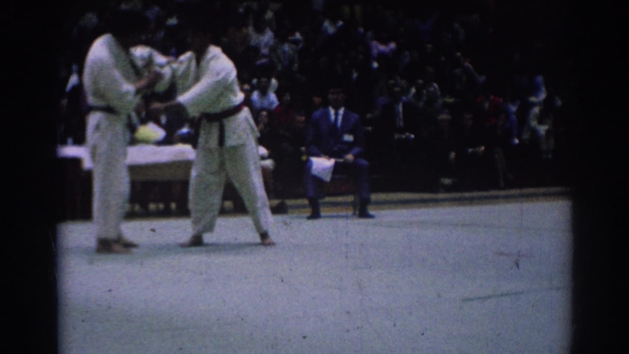 Two black-belt karate competitors in white gi continue a close hold on the tournament mat, with a seated crowd behind them.