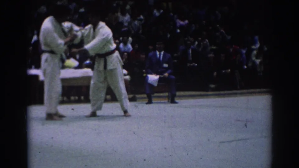Two black-belt karate competitors in white gi continue a close hold on the tournament mat, with a seated crowd behind them.